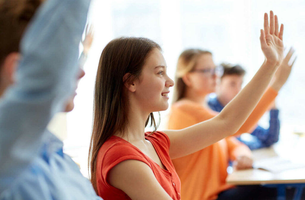 A young, smiling female student raising her hand in class surrounded by her classmates.