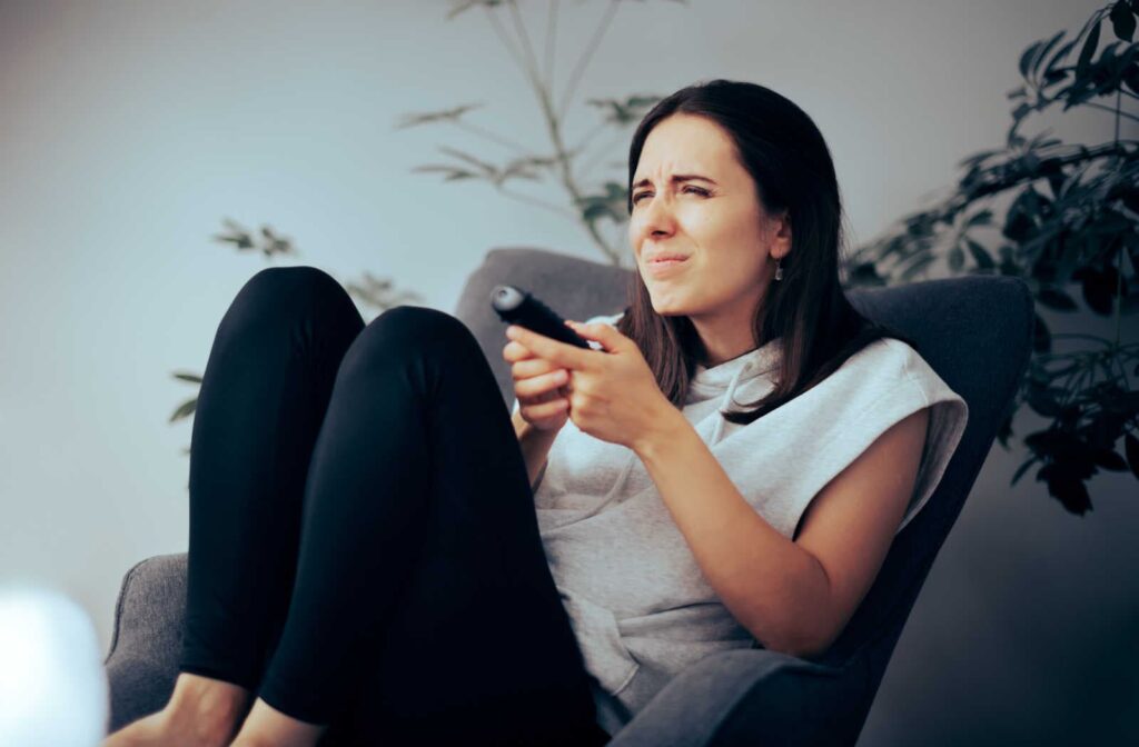 A woman sitting in a chair while holding a TV remote, squinting to try and see the TV in the distance to change the channel