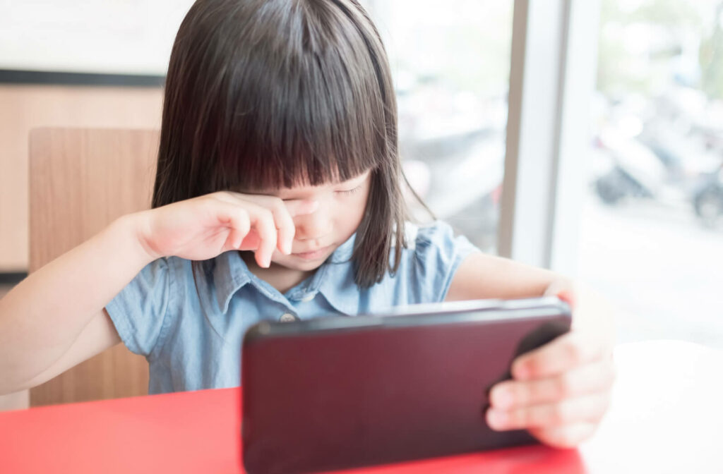 A child holding a smartphone on a table using her left hand while rubbing her right eye with her other hand.