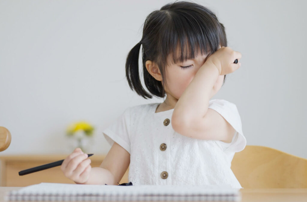 A young girl rubbing her eyes while sitting at a table trying to draw.