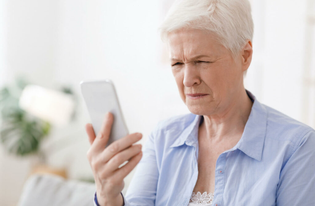An older adult woman squinting her eyes to see more clearly while holding a phone in her left hand.