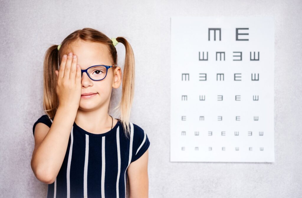 A young girl wearing glasses and covering one eye while trying to read an eye chart at an eye exam with her eye doctor.