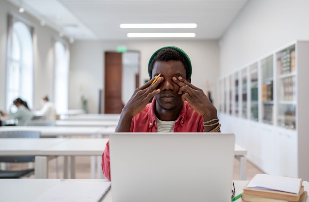 A young adult with digital eye strain rubs their eyes in front of a white laptop in a bright classroom.
