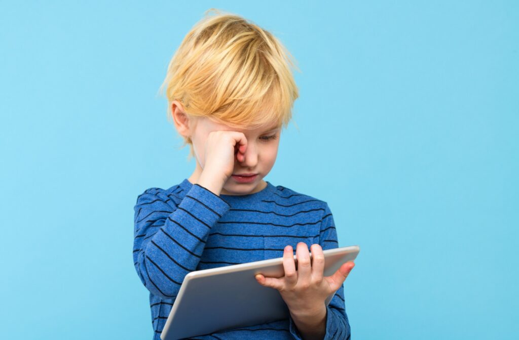 A child rubs their eyes as they spend time focusing on their tablet screen.